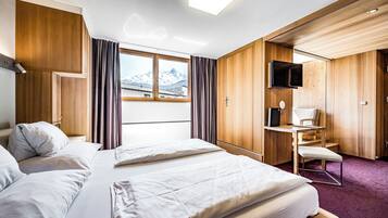Ein Hotelzimmer mit zwei Betten, einem Schreibtisch, einem Sessel, einem Fernseher und einem Fenster mit Blick auf die Berge.