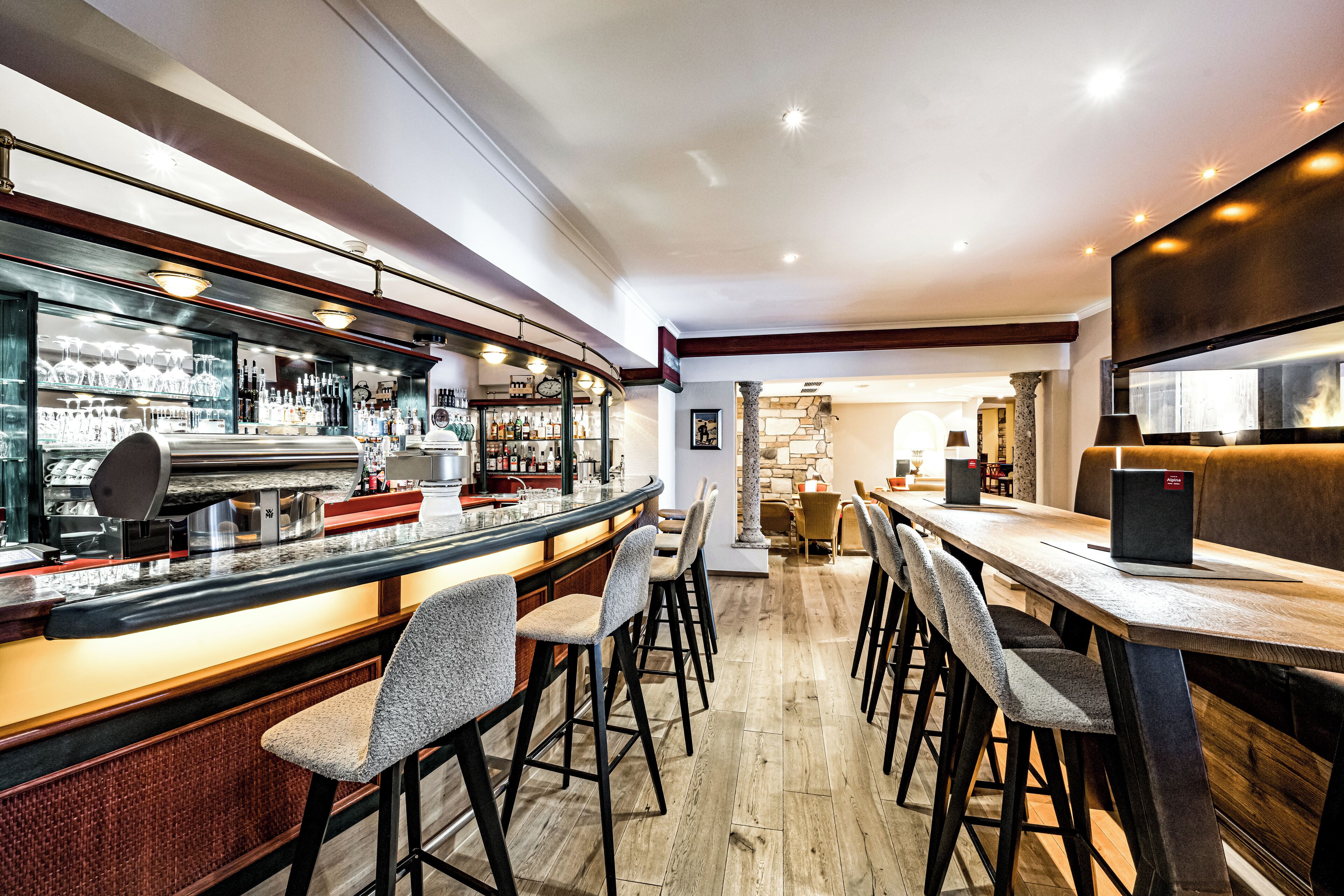 A bar area with stools, a counter with glasses and bottles, and a wooden table with chairs.