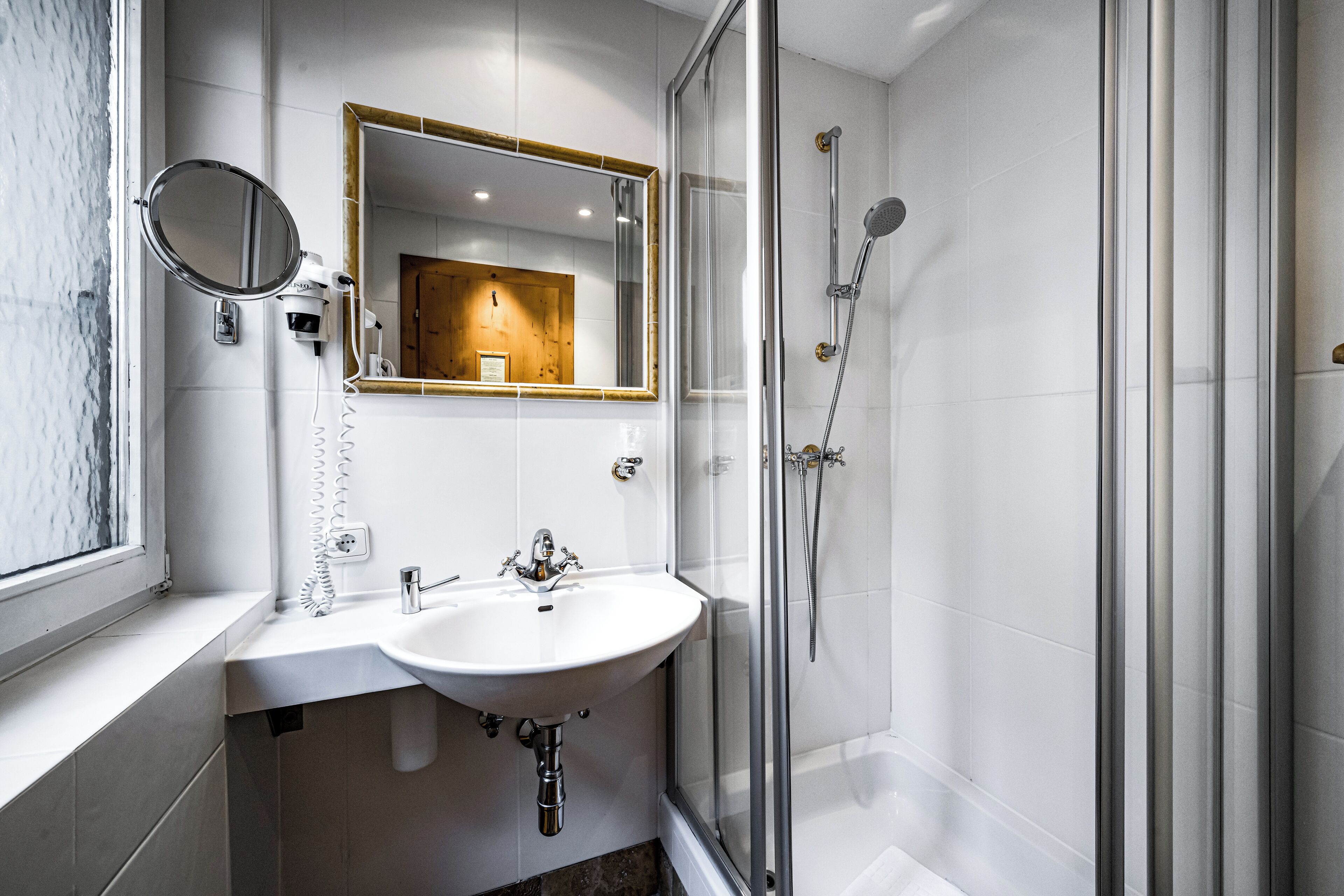 A modern bathroom with a white sink, a round mirror, a large mirror above the sink, and a glass-enclosed shower area.
