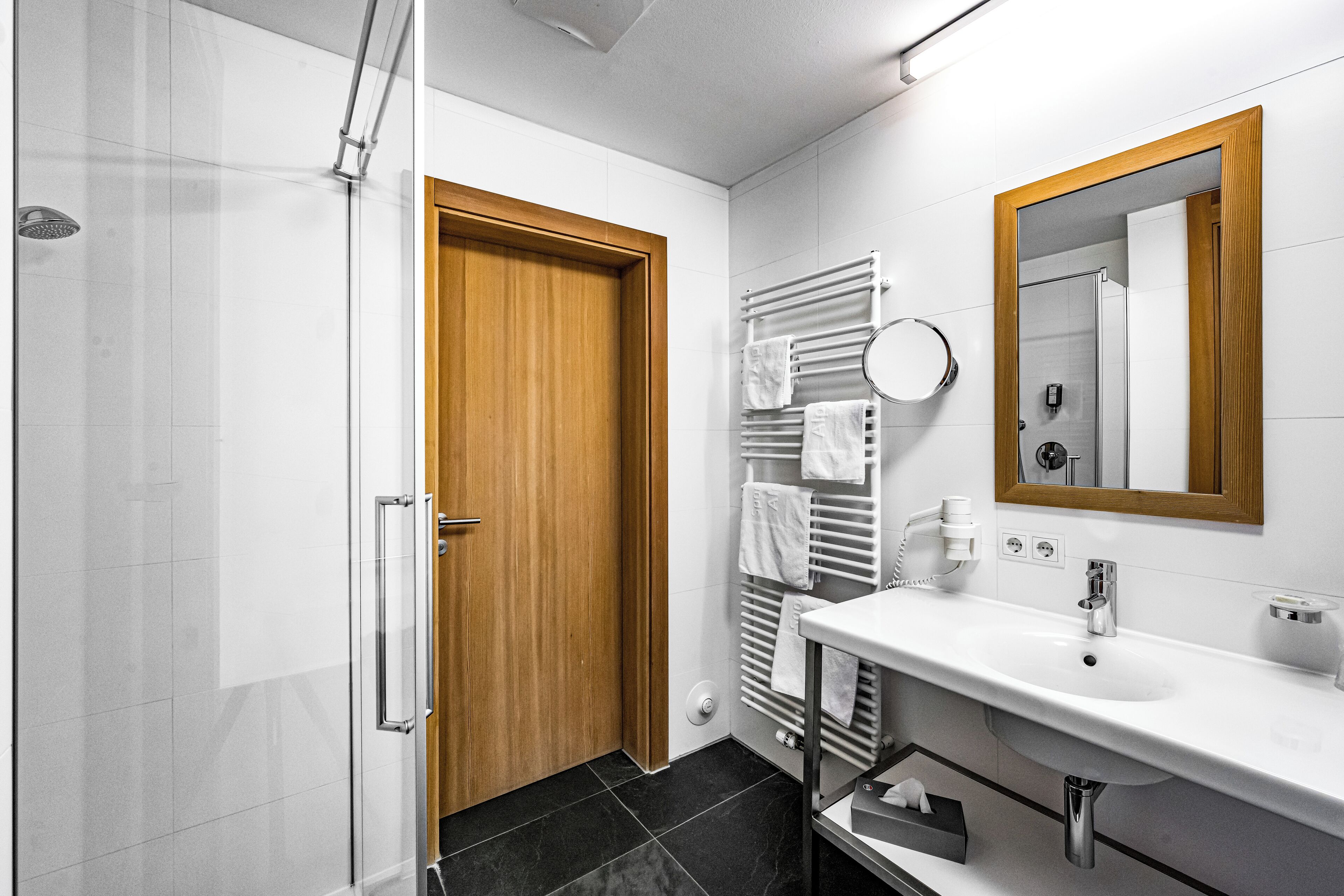 Ein modernes Badezimmer mit Dusche, Holztür, weißem Waschbecken und einem Spiegel mit Holzrahmen.