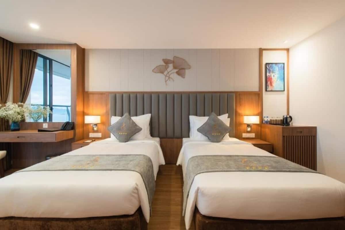 A hotel room with two beds, a wooden headboard, bedside lamps, a desk, and a window with curtains.