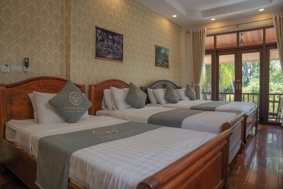 A hotel room with three wooden beds, each with a grey bedspread and pillows, and a large window with a view of greenery.