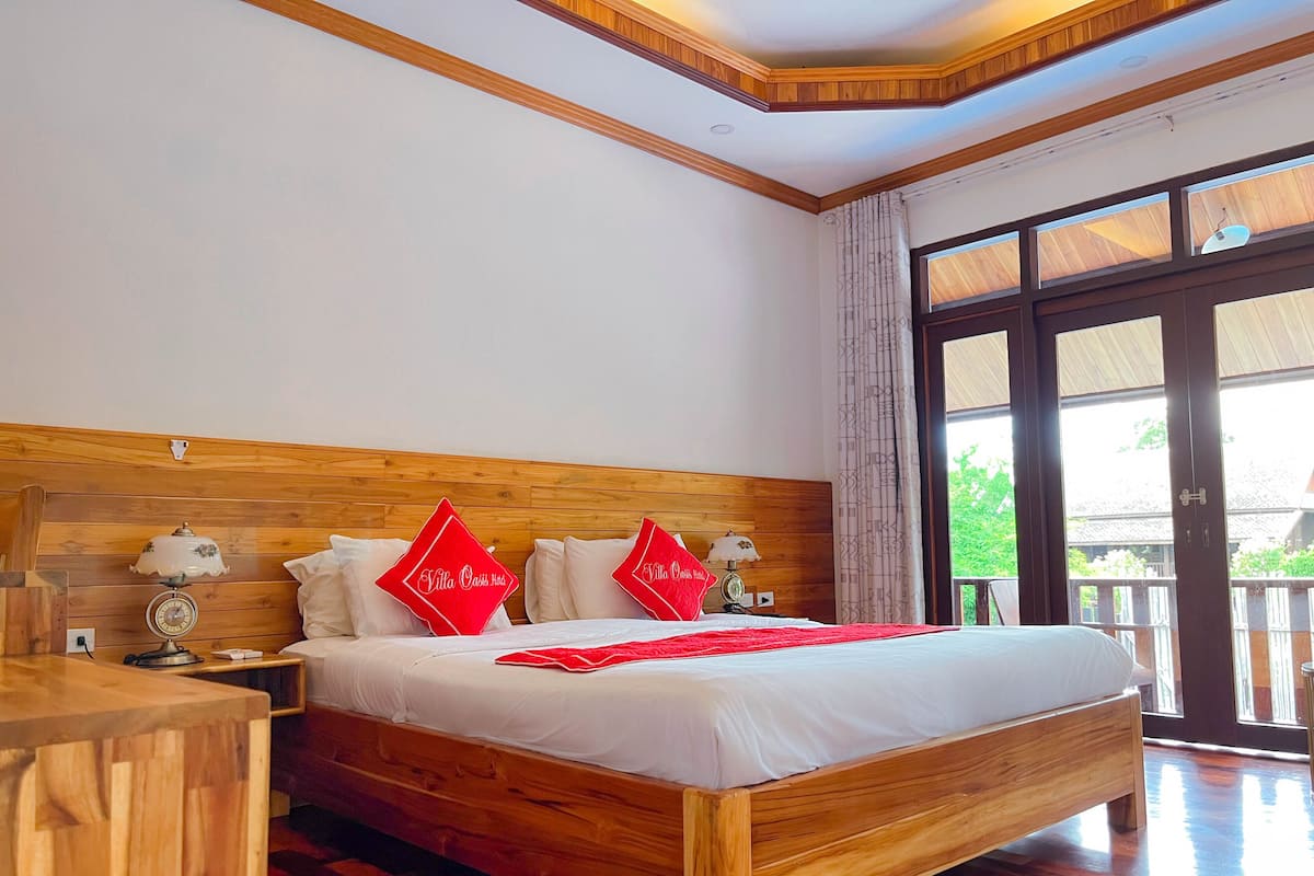 A wooden bed with a white mattress and red pillows, a bedside table with a lamp, and a large window with curtains.