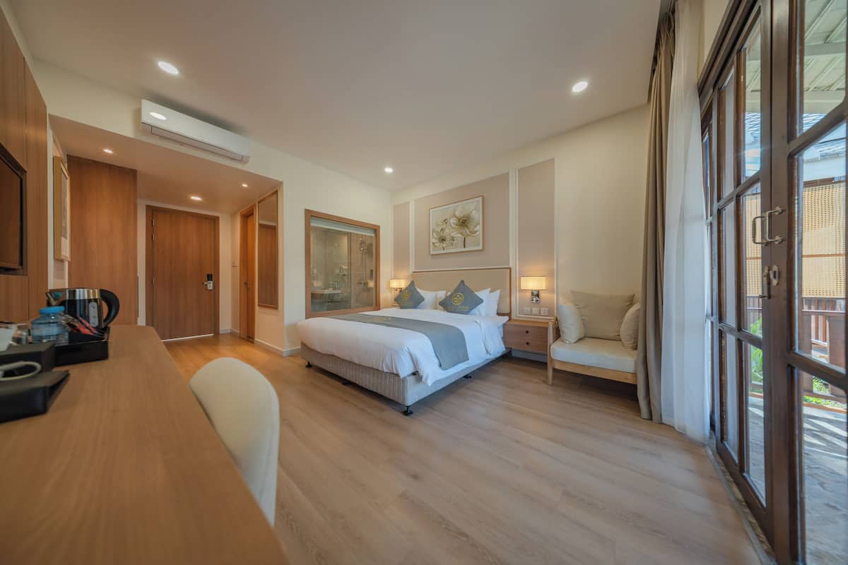 A hotel room with a wooden floor, a bed with white bedding, a desk with a chair, a bedside table with a lamp, and a large window with curtains.