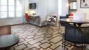 Une chambre d’hôtel équipée d’une table à manger, de chaises, d’un téléviseur fixé au mur, d’une moquette à motifs et d’une fenêtre avec des rideaux.