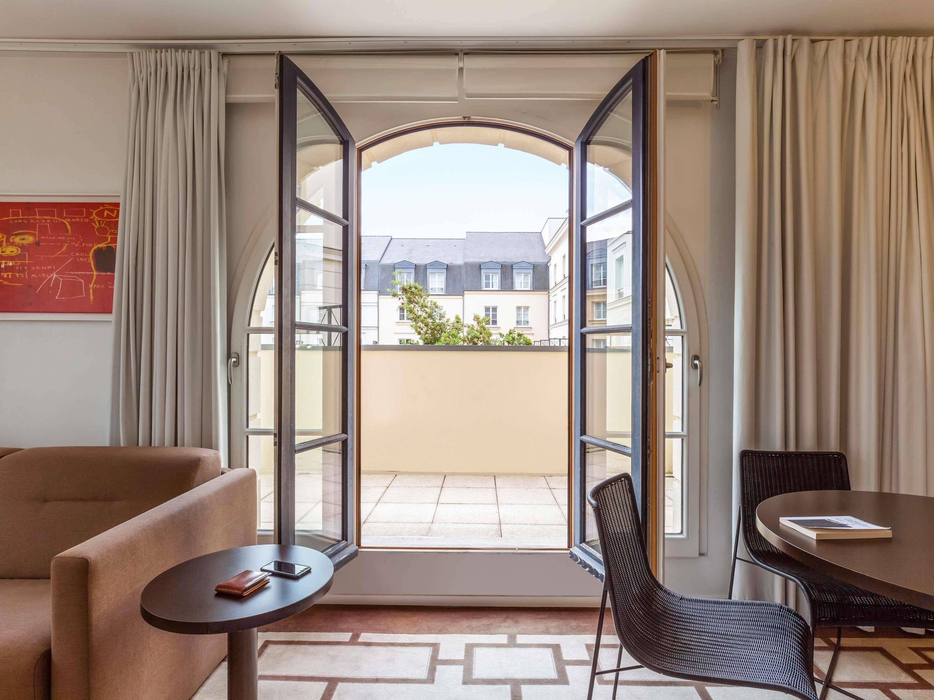 A hotel room with a large arched window, a sofa, a small table, a chair, and a dining table.