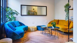 Ein modernes Wohnzimmer mit blauem Sessel, gelbem Sofa und rundem Couchtisch. Zwei kleine Hocker, ein großer Spiegel an der Wand und ein Blumentopf auf einem Metallständer sind ebenfalls vorhanden.