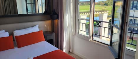 A hotel room with a large bed, a nightstand, a mirror, and a window with a view of a green landscape.