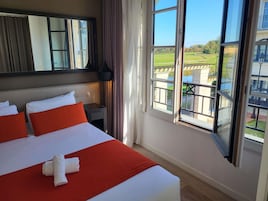 Habitación de hotel con una cama grande, una mesita de noche, un espejo y una ventana con vistas a un paisaje verde.