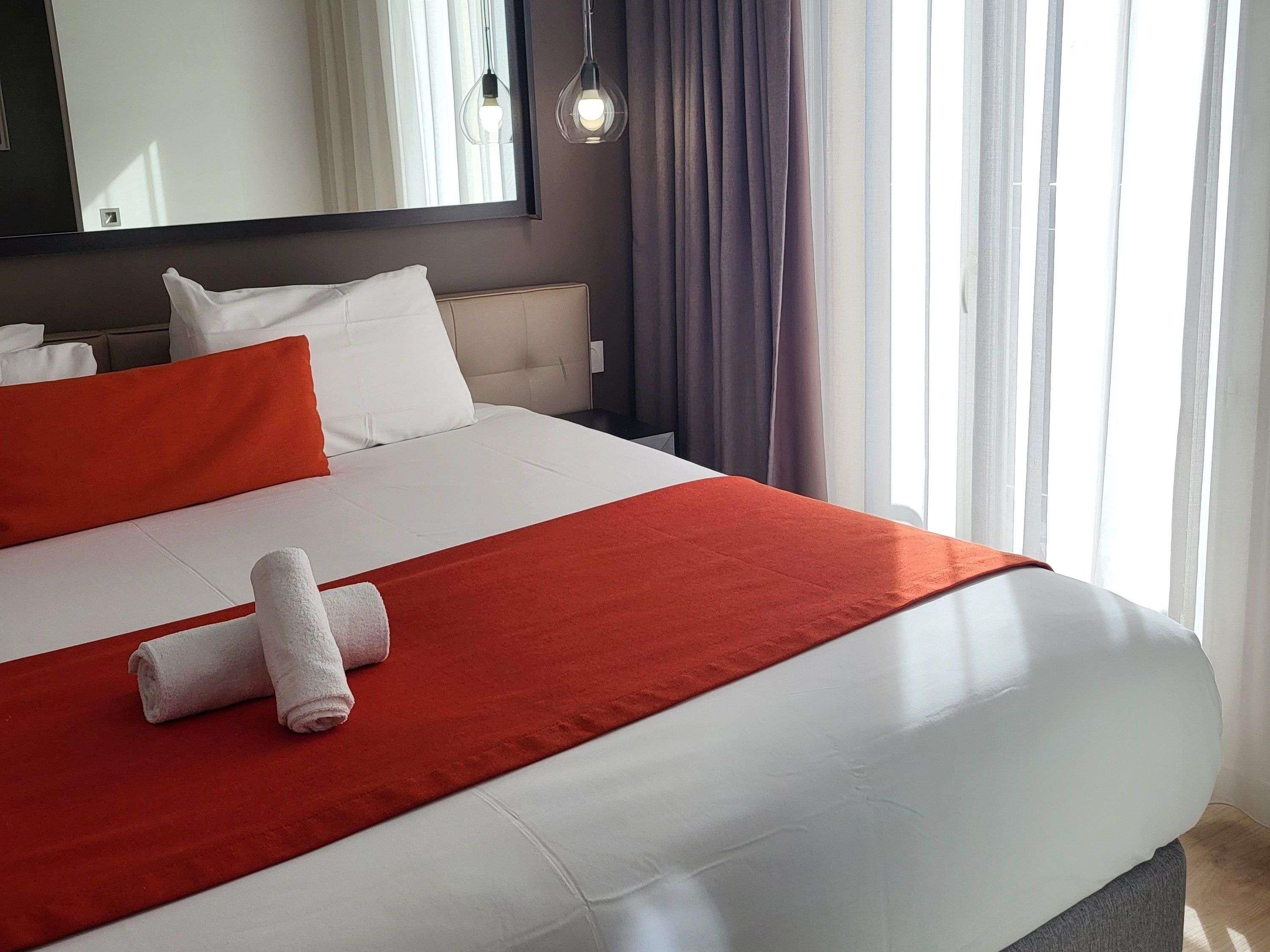 Une chambre d’hôtel avec un lit, du linge de lit blanc, un plaid rouge et une serviette enroulée.