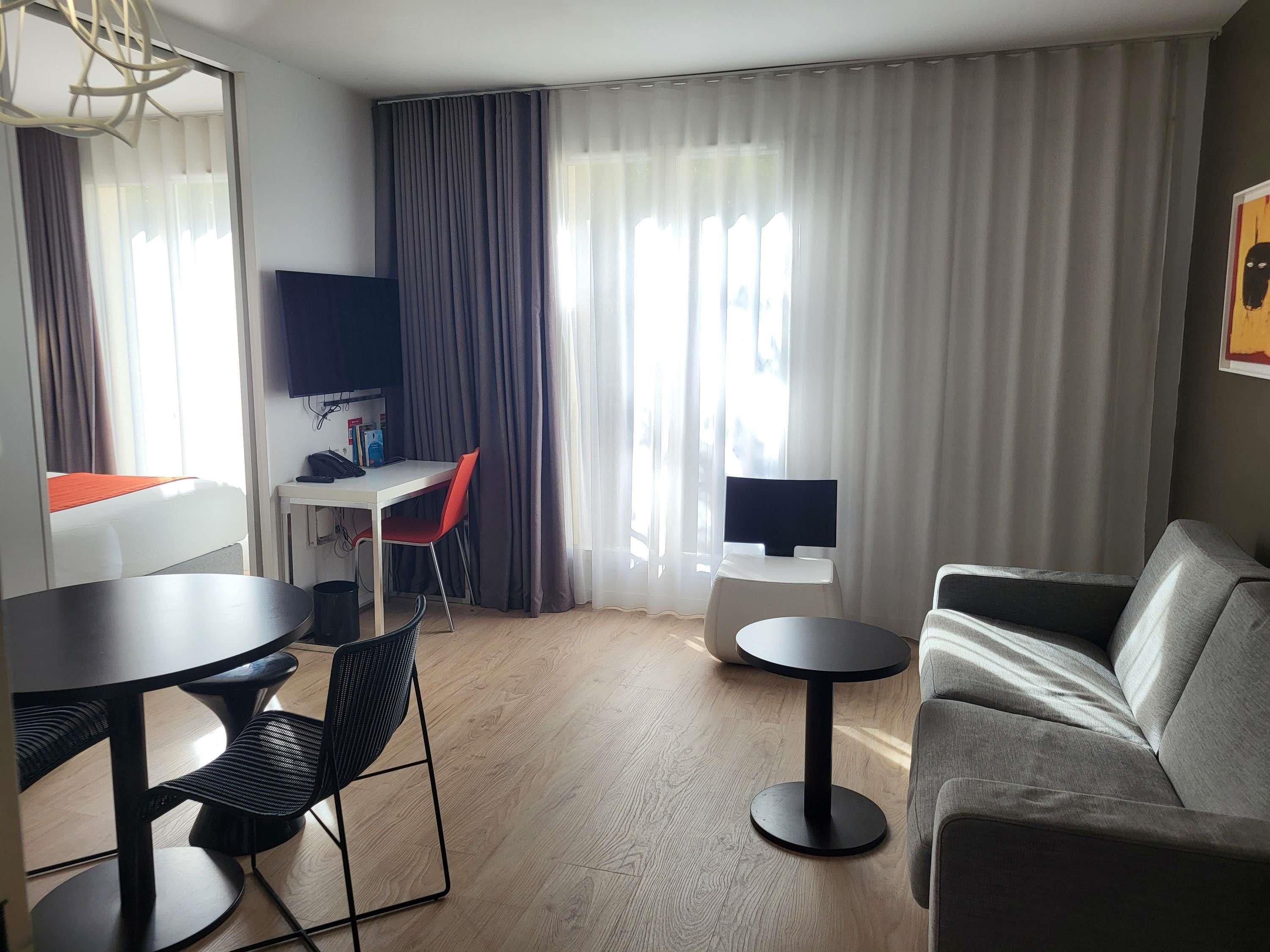A modern hotel room with a grey sofa, a round dining table with chairs, a small white desk with a chair, a flat-screen TV mounted on the wall, and a large window with grey curtains.