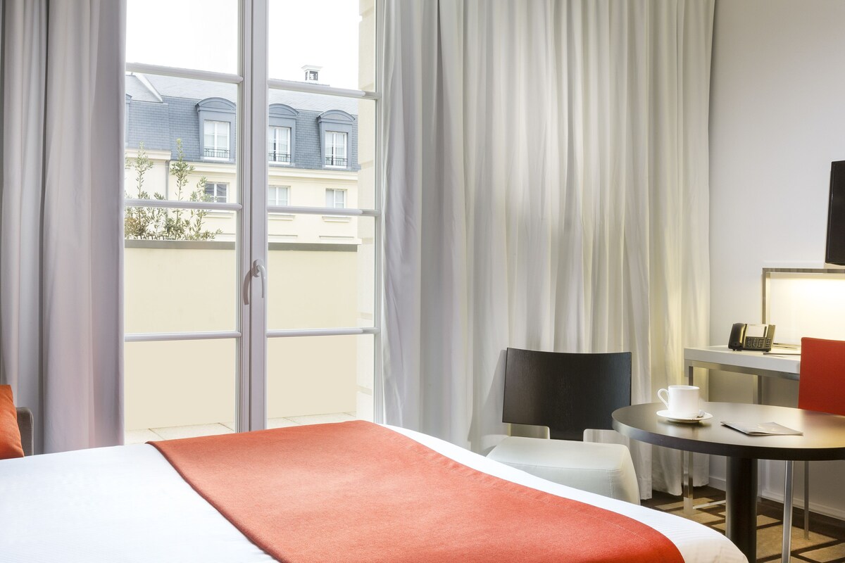 Quarto de hotel com cama, mesa com xícara e pires, cadeira e televisão.