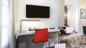 Ein Hotelzimmer mit Schreibtisch, einem roten Sessel, einem Bett mit Kuhmusterdecke, einem an der Wand montierten Fernseher und einem gemusterten Teppich.