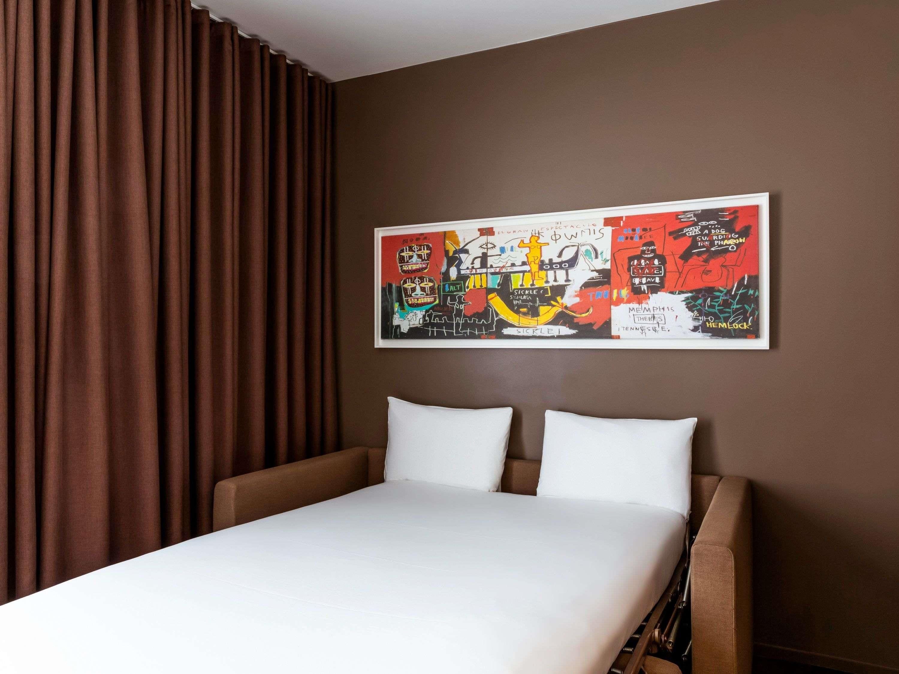 A hotel room with a bed, brown curtains, and a colorful abstract painting on the wall.