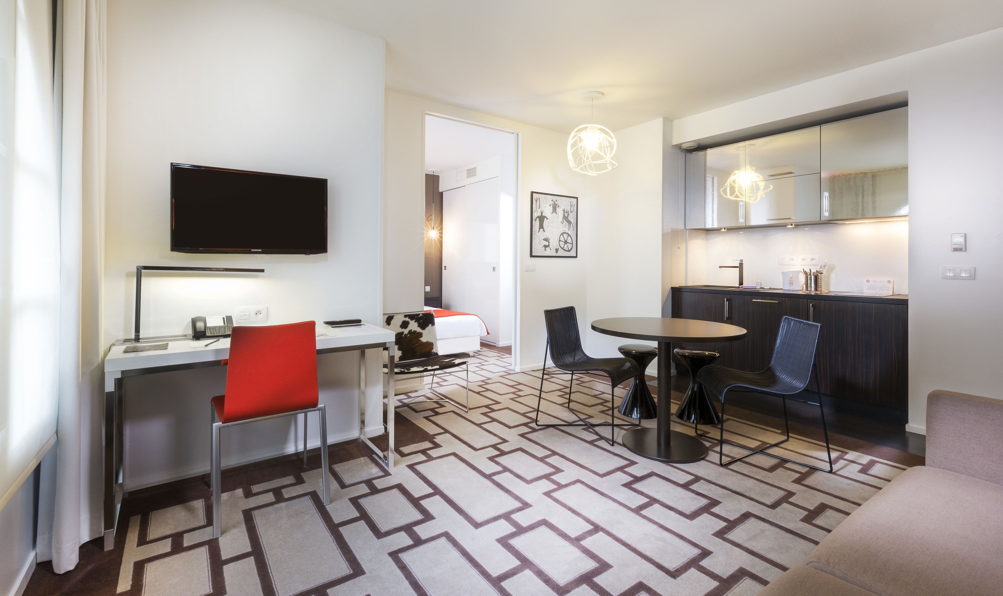 Ett modernt hotellrum med en platt-TV, ett skrivbord med en stol, en matplats med bord och stolar, ett kök med en spegel och en mönstrad matta.