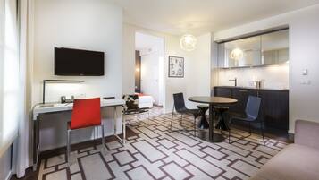 A modern hotel room with a flat-screen TV, a desk with a chair, a dining area with a table and chairs, a kitchen with a mirror, and a patterned carpet.