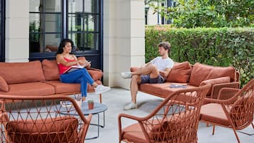 An outdoor seating area with wicker furniture, a glass table, and a building with large windows in the background.