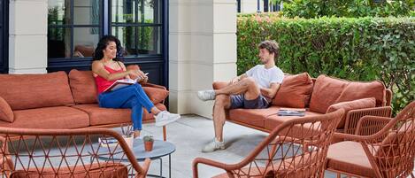 An outdoor seating area with wicker furniture, a glass table, and a building with large windows in the background.