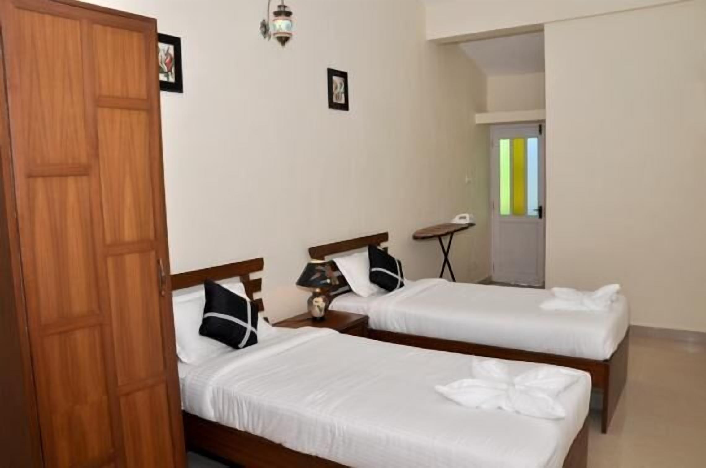 A hotel room with two beds, a wooden wardrobe, a small table, and a wall-mounted lamp.
