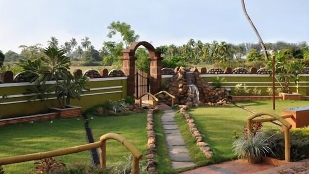 A garden with a stone pathway leading to a gate, surrounded by greenery and palm trees. - A's Holiday Beach Resort (Betalbatim)