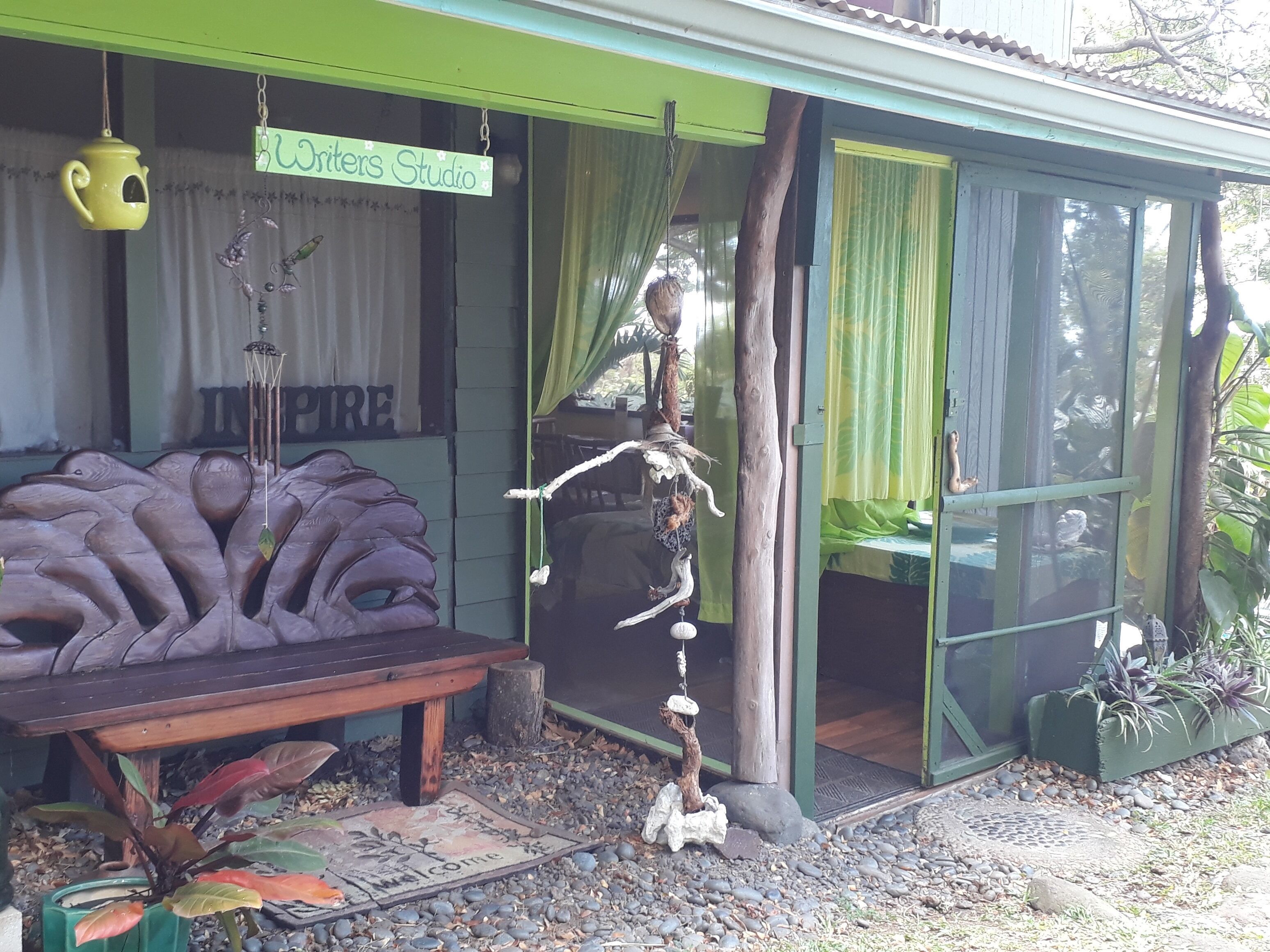 A studio with a green exterior, a wooden bench, a welcome mat, and a hanging bird feeder.