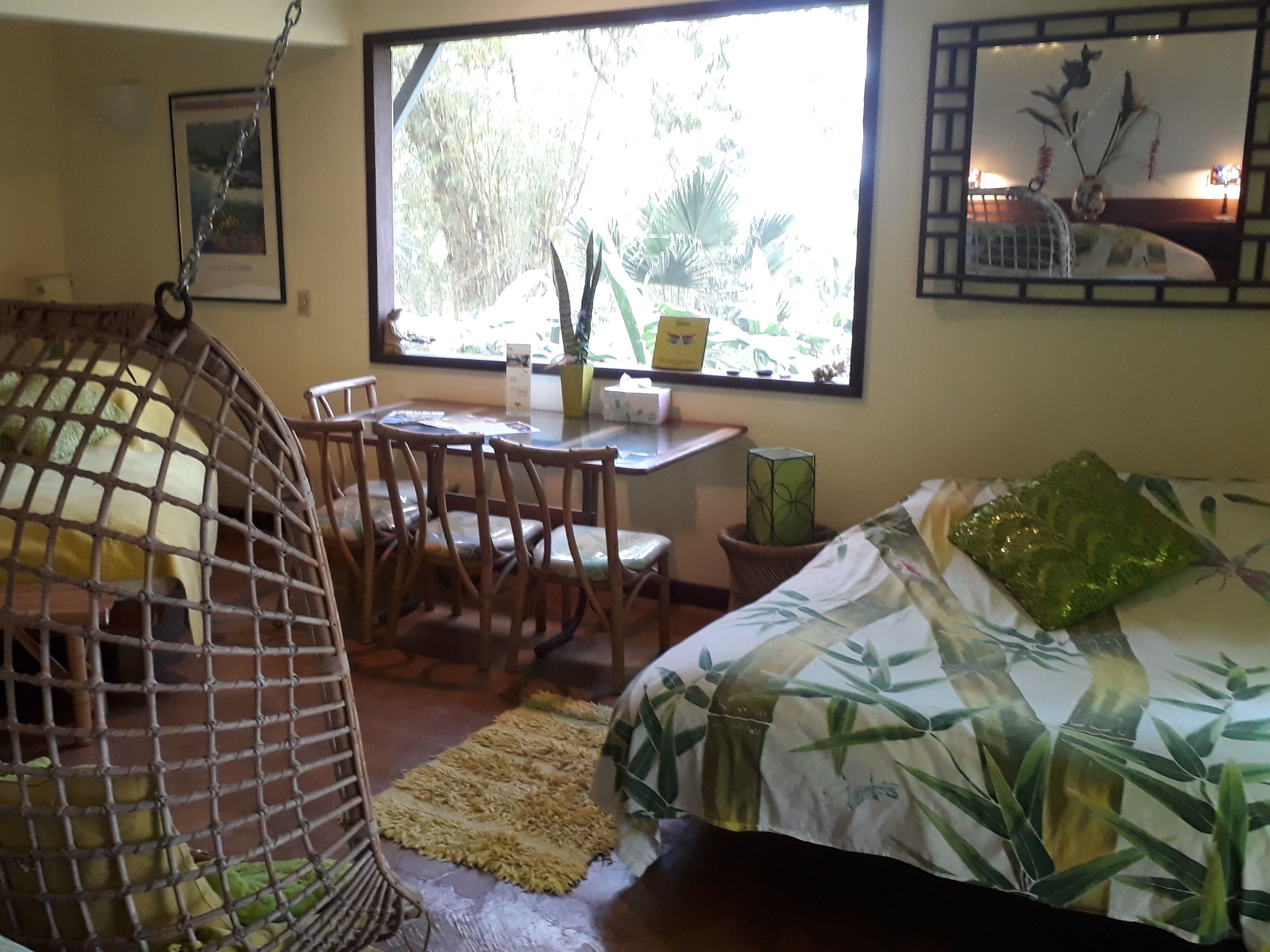 Une pièce comprenant un lit, un fauteuil suspendu, une table avec des chaises, une fenêtre donnant sur des arbres, et une petite table sur laquelle se trouvent un vase et une plante.
