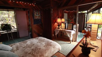 Une chambre confortable avec un lit, une table de chevet et un bureau avec des lampes. Il y a une fenêtre avec des rideaux et une vue sur la verdure à l’extérieur.