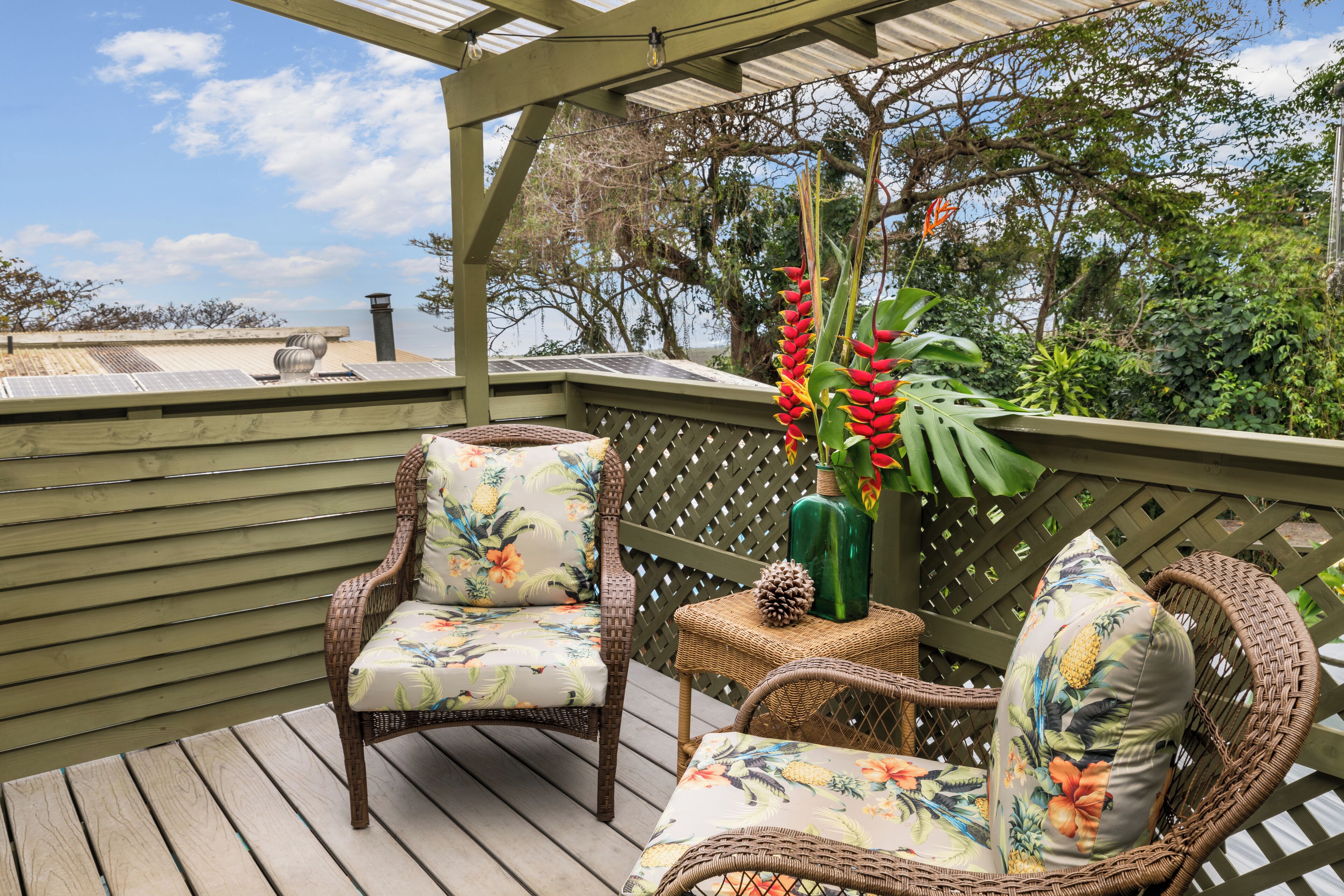 A wooden deck with wicker chairs and a table, surrounded by lush greenery and a view of the sky.