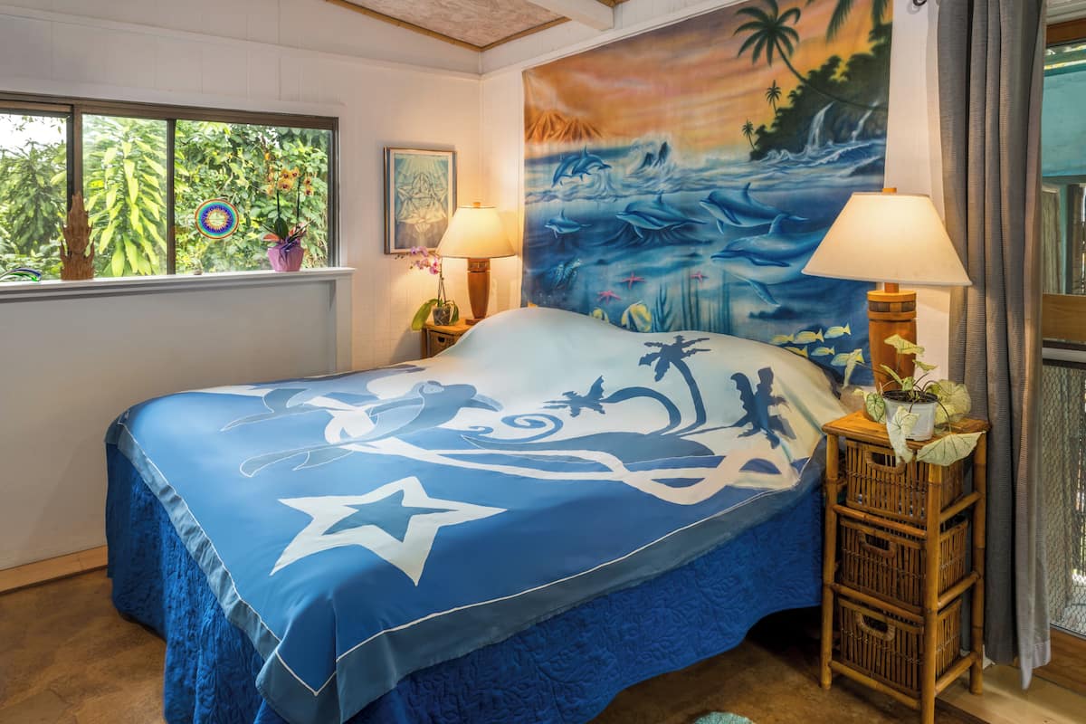 Een bed met een tropisch deken in blauw en wit, twee bedlampjes, een muurschildering van een tropisch landschap en een raam met uitzicht op groen.