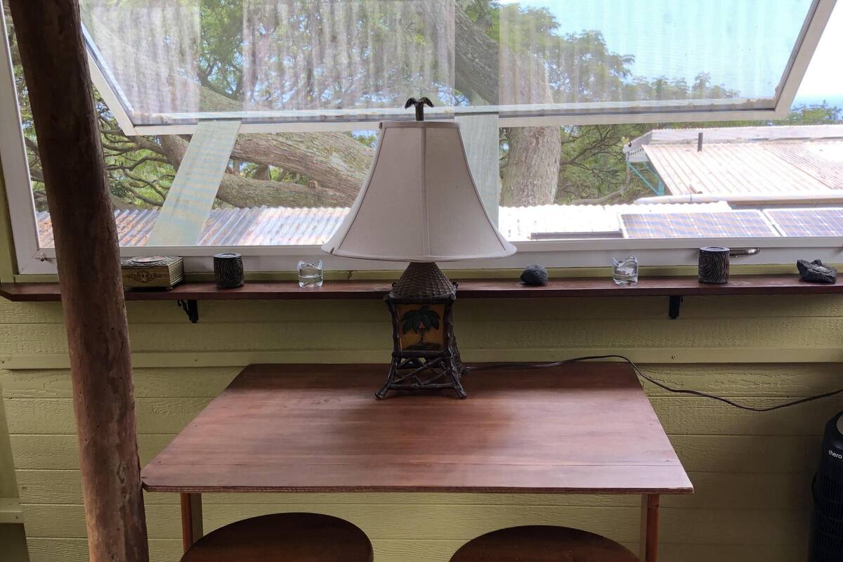 Een kamer met een houten tafel en twee krukken, een raam met uitzicht op bomen en een dak, en een lamp op de tafel.