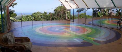 Una cancha deportiva interior con un diseño circular de colores, rodeada por un recinto de vidrio y sillas de mimbre.