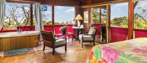 A room with a wooden ceiling, a large bed with a floral bedspread, two chairs, a small table with a lamp, a red chair, and a view of trees and sky through large windows.