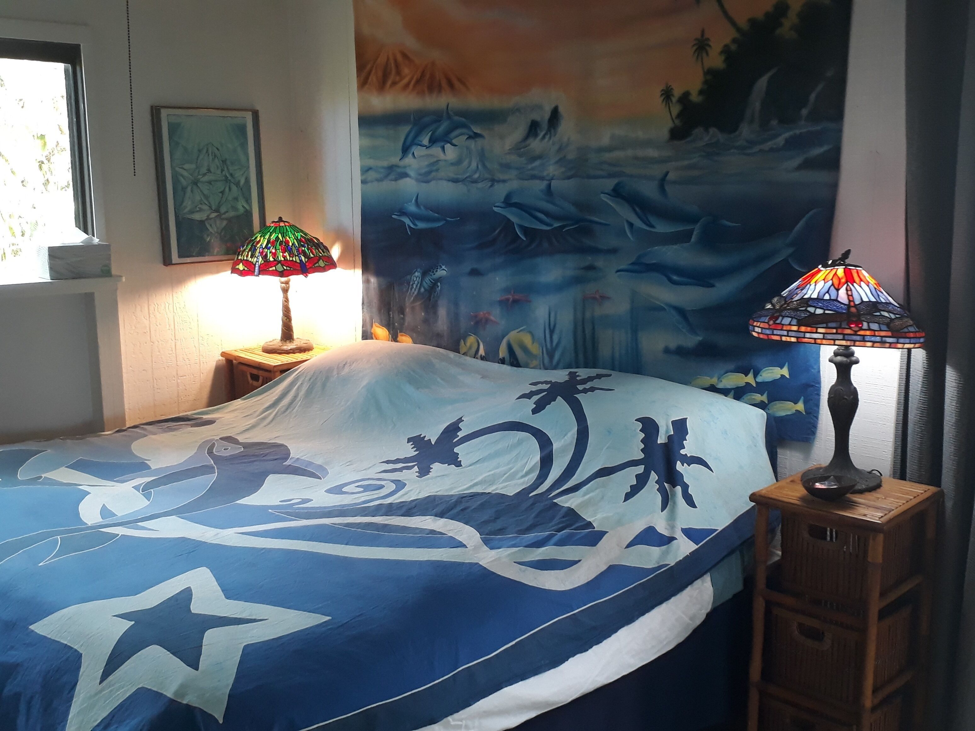Een slaapkamer met een bed, voorzien van een blauw-witte bedspreide in nautische stijl, twee wandlampen en een groot wandkleed waarop dolfijnen en een tropisch landschap afgebeeld zijn.