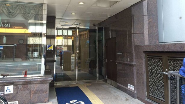 Entrada de hotel con puerta de vidrio, felpudo azul y señal de accesibilidad para sillas de ruedas.
