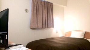 Una habitación de hotel con una cama, un escritorio con computadora y controles remotos, una ventana con cortinas y una lámpara fijada a la pared.