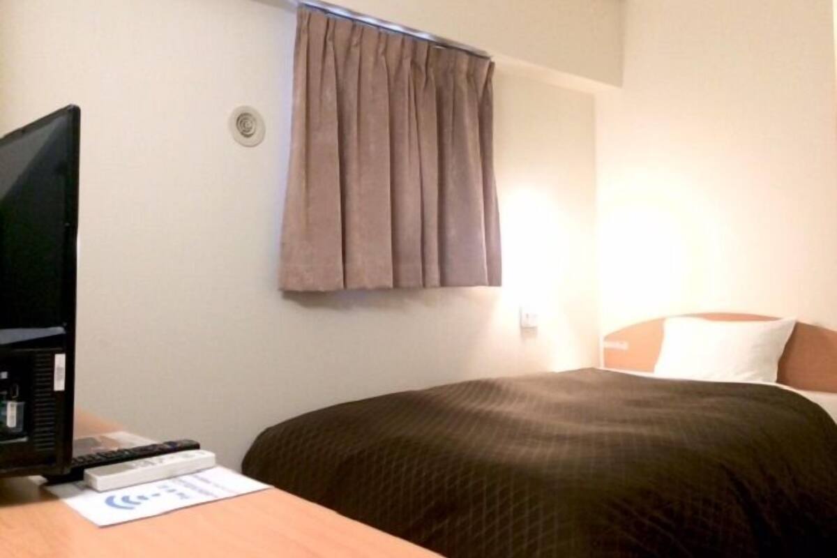 A hotel room with a bed, a desk with a computer and remote controls, a window with curtains, and a wall-mounted light.