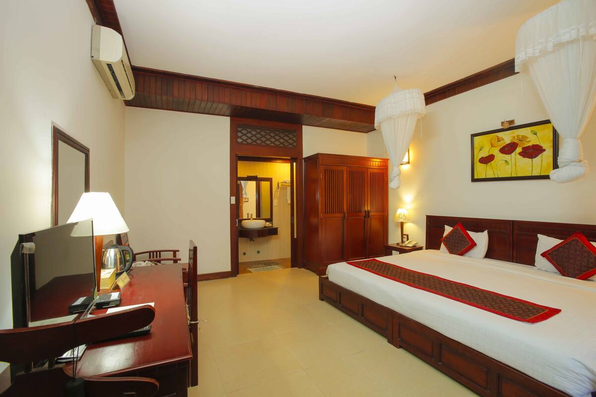 A hotel room with a bed, bedside table, lamp, desk with chair, and a bathroom visible through an open door.