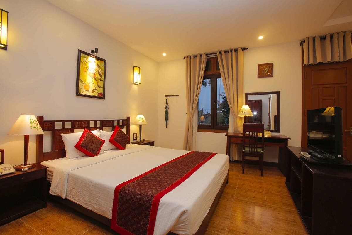 A hotel room with a large bed, bedside lamps, a desk with a chair, a television, and a window with curtains.