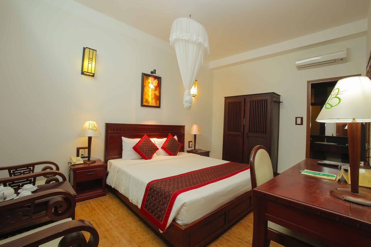 A hotel room with a bed, bedside tables, a desk, a chair, a wardrobe, and a hanging light fixture.