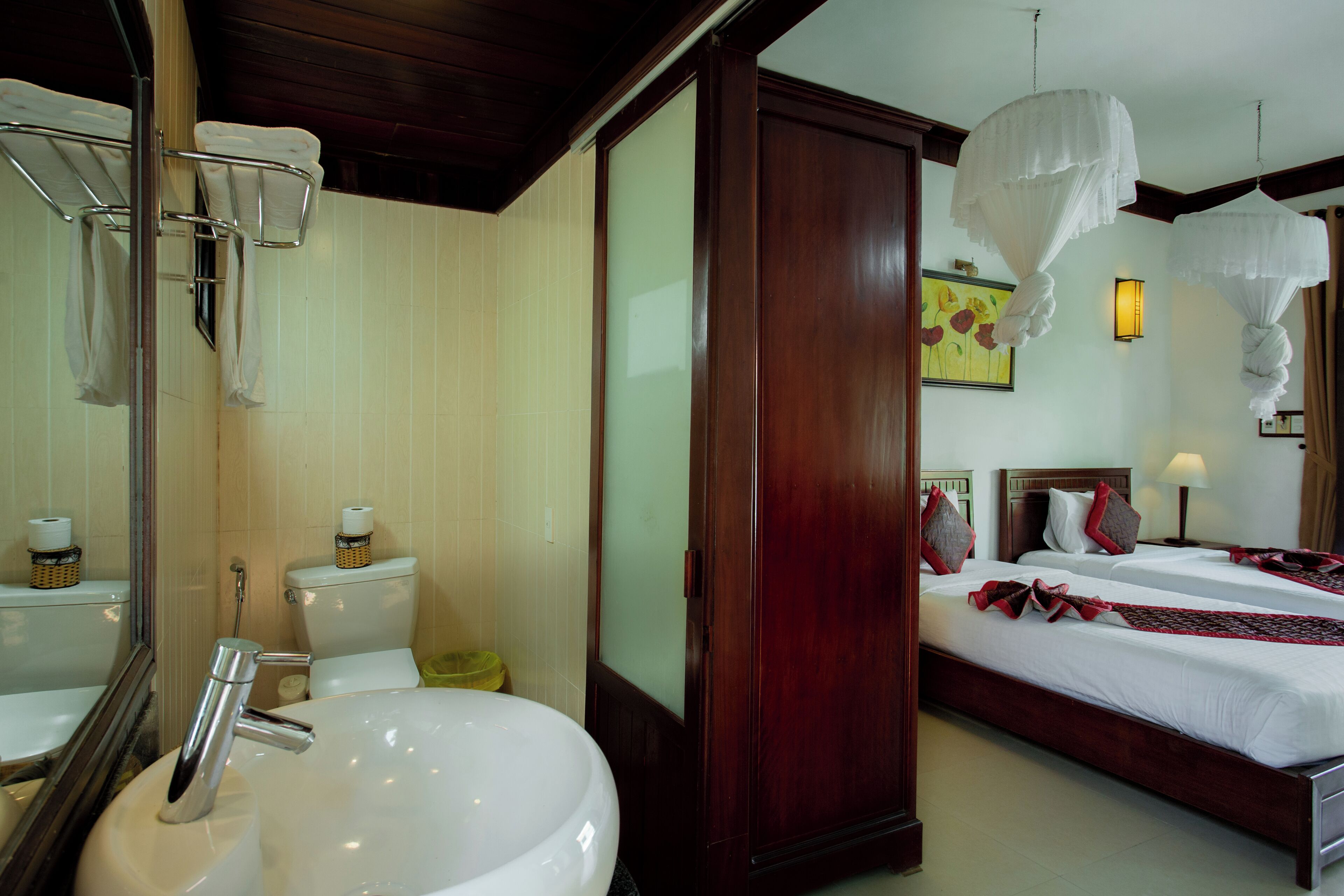 A hotel room with a large white sink, a wooden wardrobe, two beds with white bedding and red accents, and a painting on the wall.
