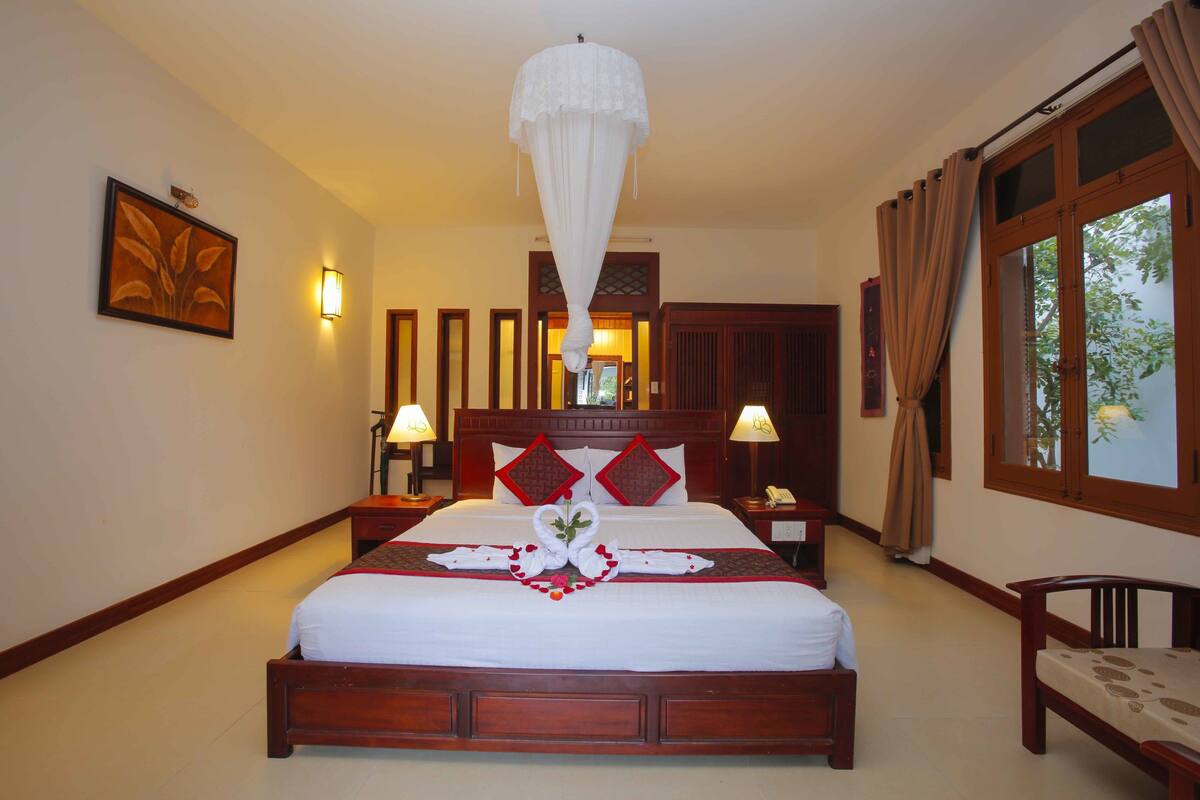 A hotel room with a large bed, wooden headboard, two bedside lamps, a nightstand, and a window with curtains.