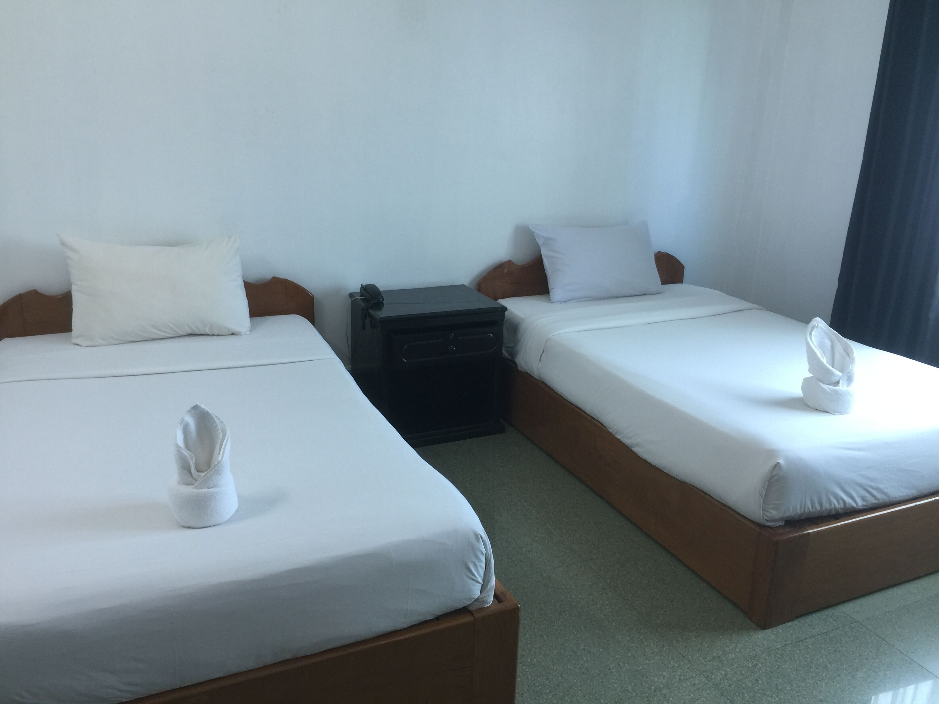 Habitación de hotel con dos camas, cada una con ropa blanca y almohadas, una mesita de noche pequeña con un teléfono, y una cama bien hecha con una toalla.
