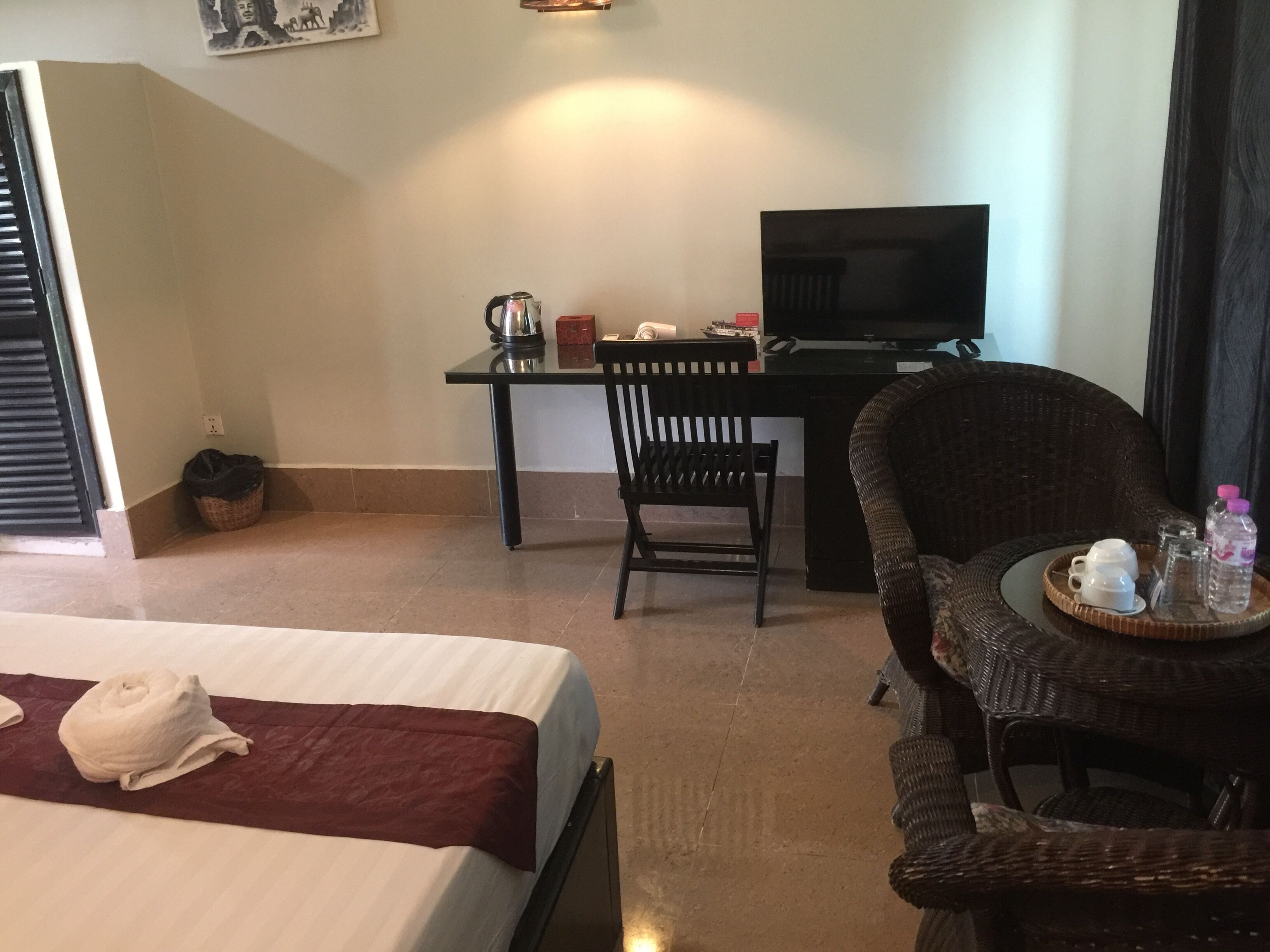 A hotel room with a bed, a desk with a television, a wicker chair, and a small table with a tray of items.