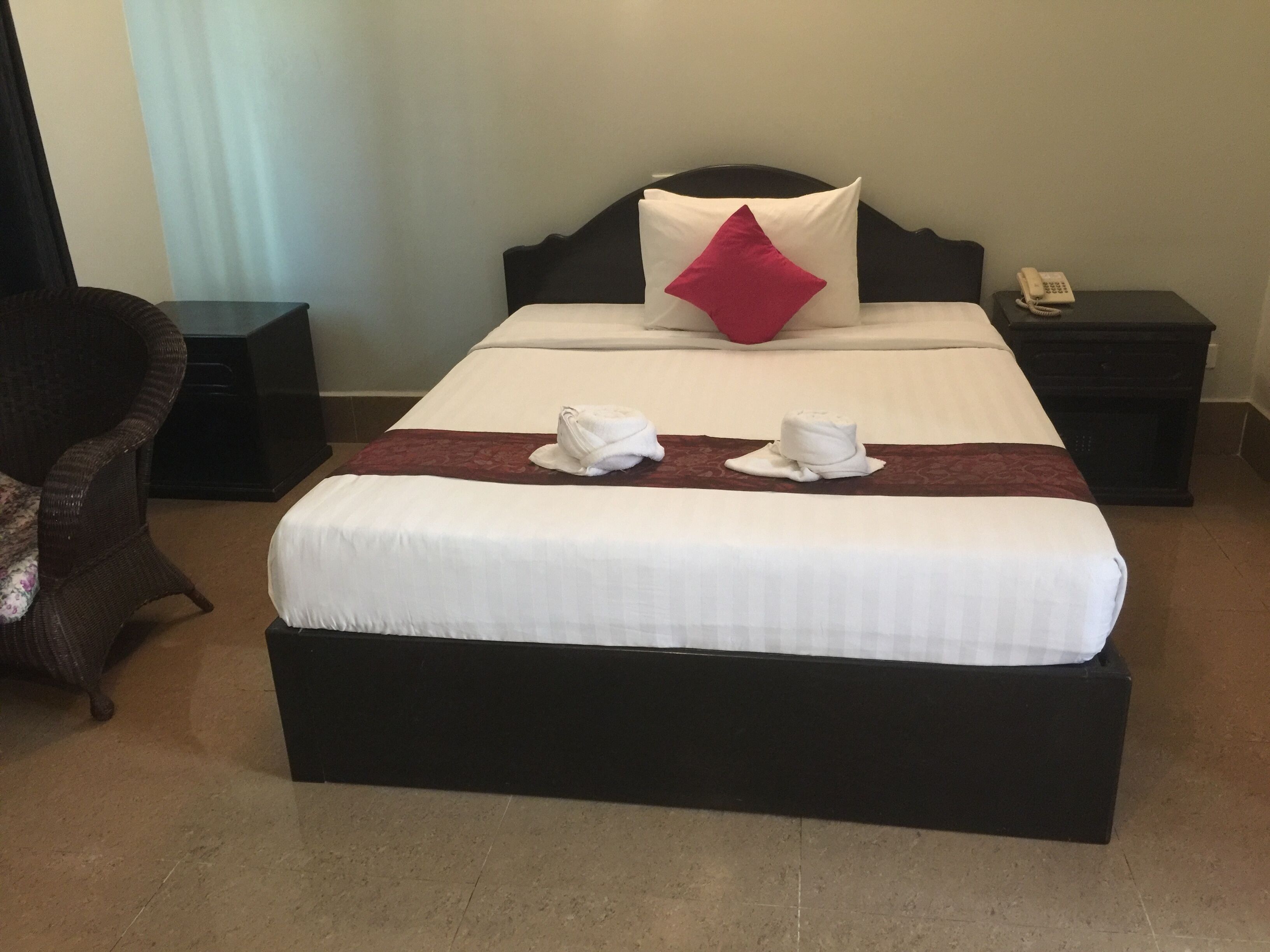 Camera d'albergo con un letto grande, un cuscino rosso, due asciugamani bianchi, un telefono sul comodino e una sedia in vimini.