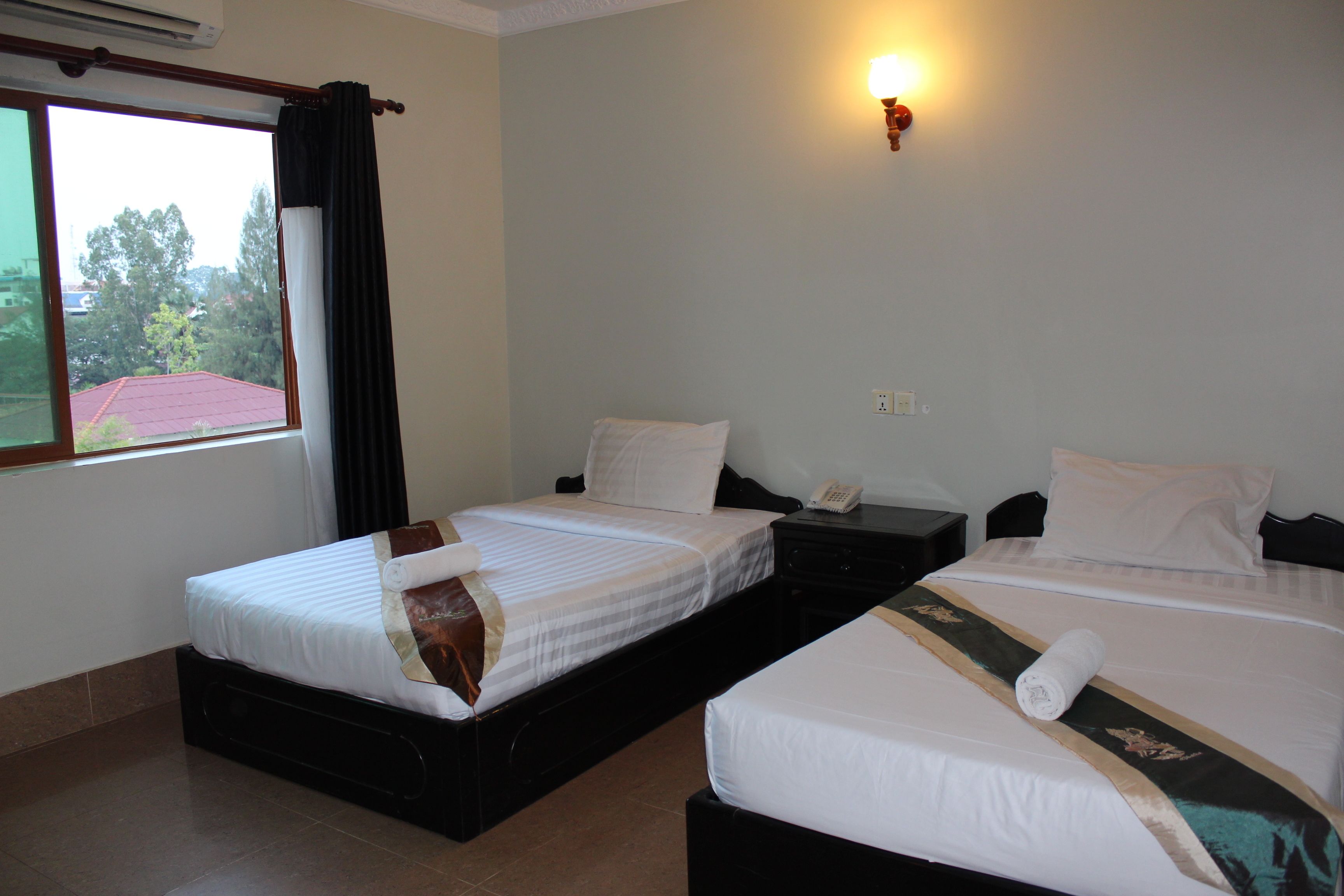 A hotel room with two beds, a window with curtains, a bedside table with a phone, and a wall-mounted light.