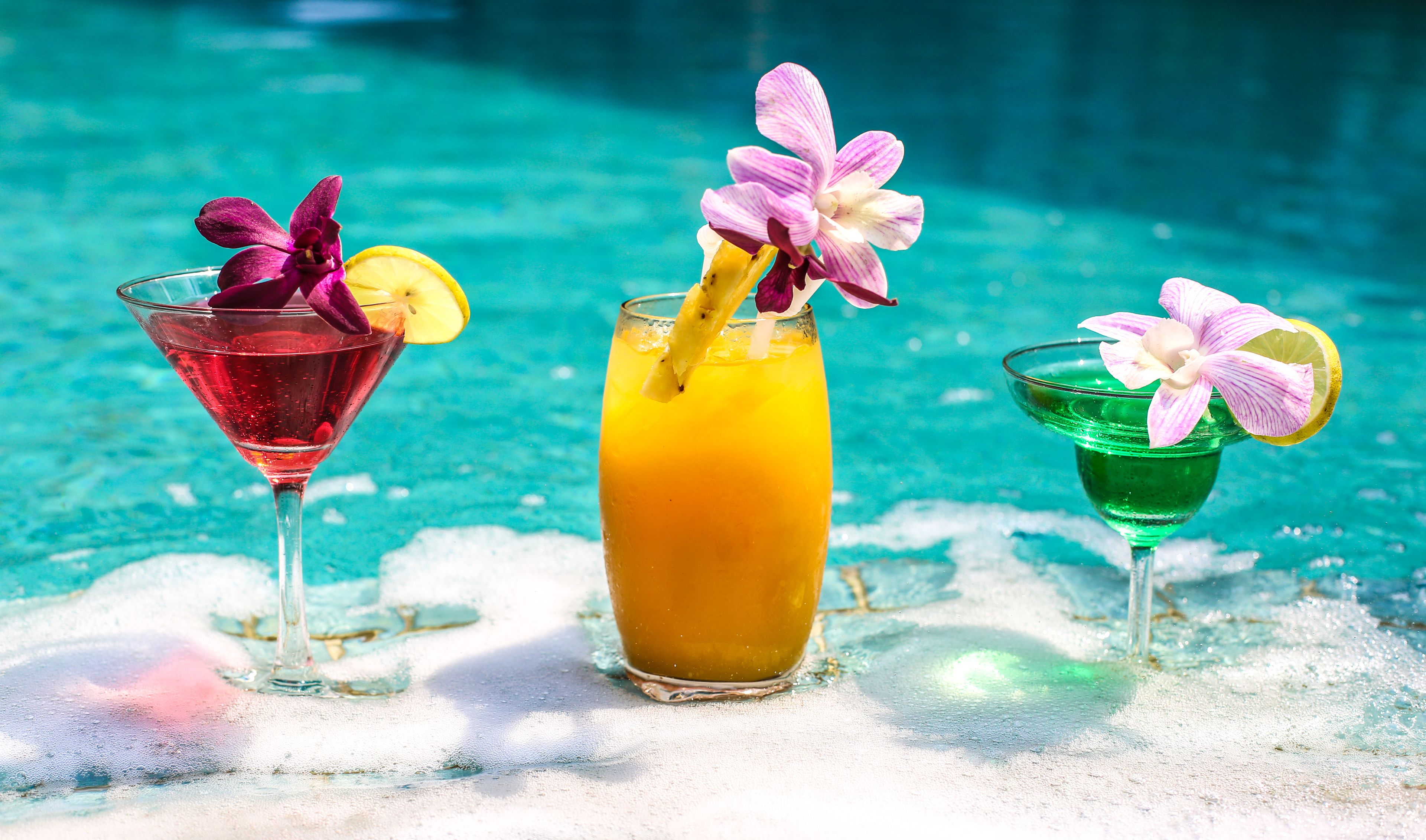 Three cocktails with flowers and fruit garnishes by a pool.