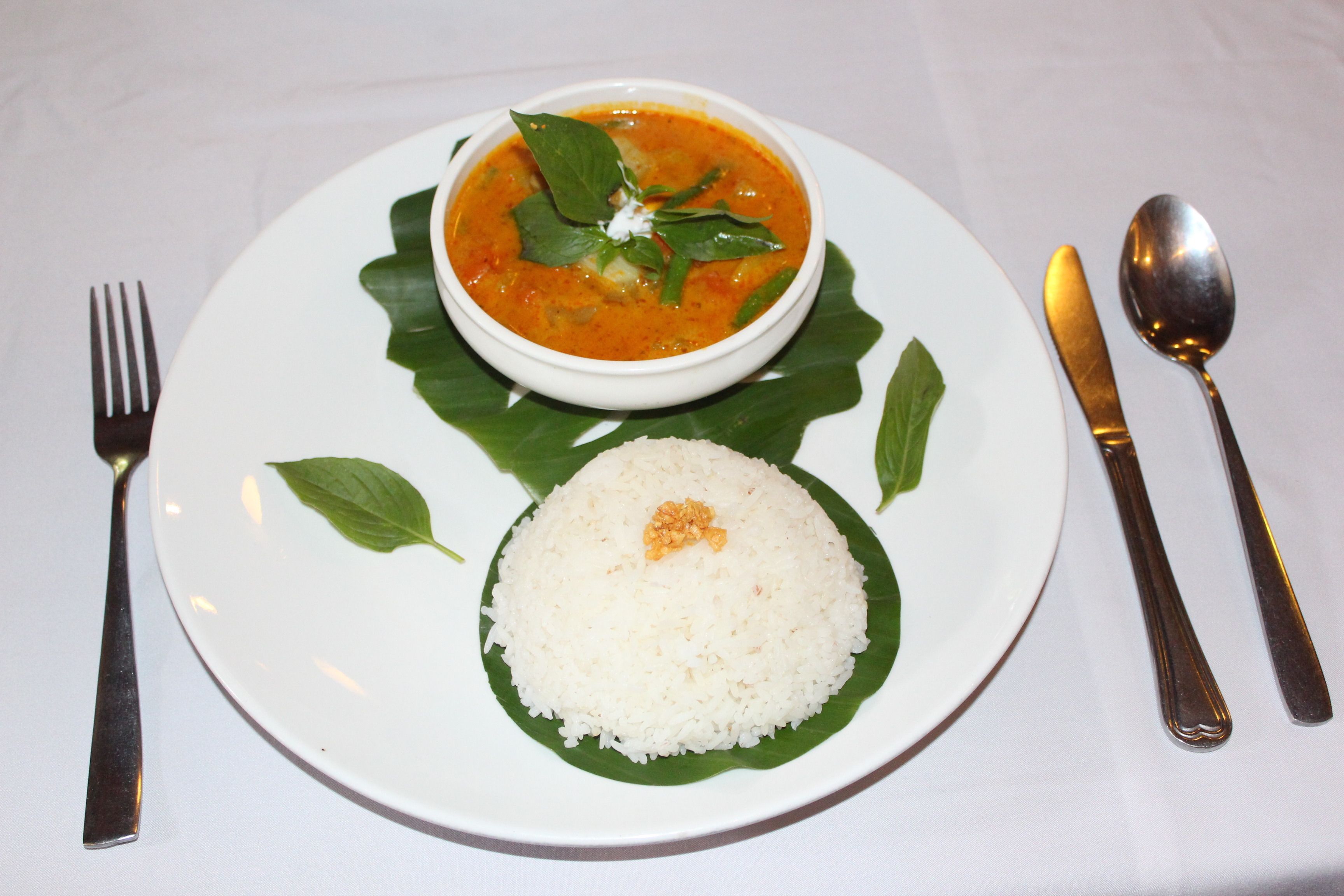 A plate with white rice and a bowl of soup, accompanied by a fork, knife, and spoon on a white tablecloth.
