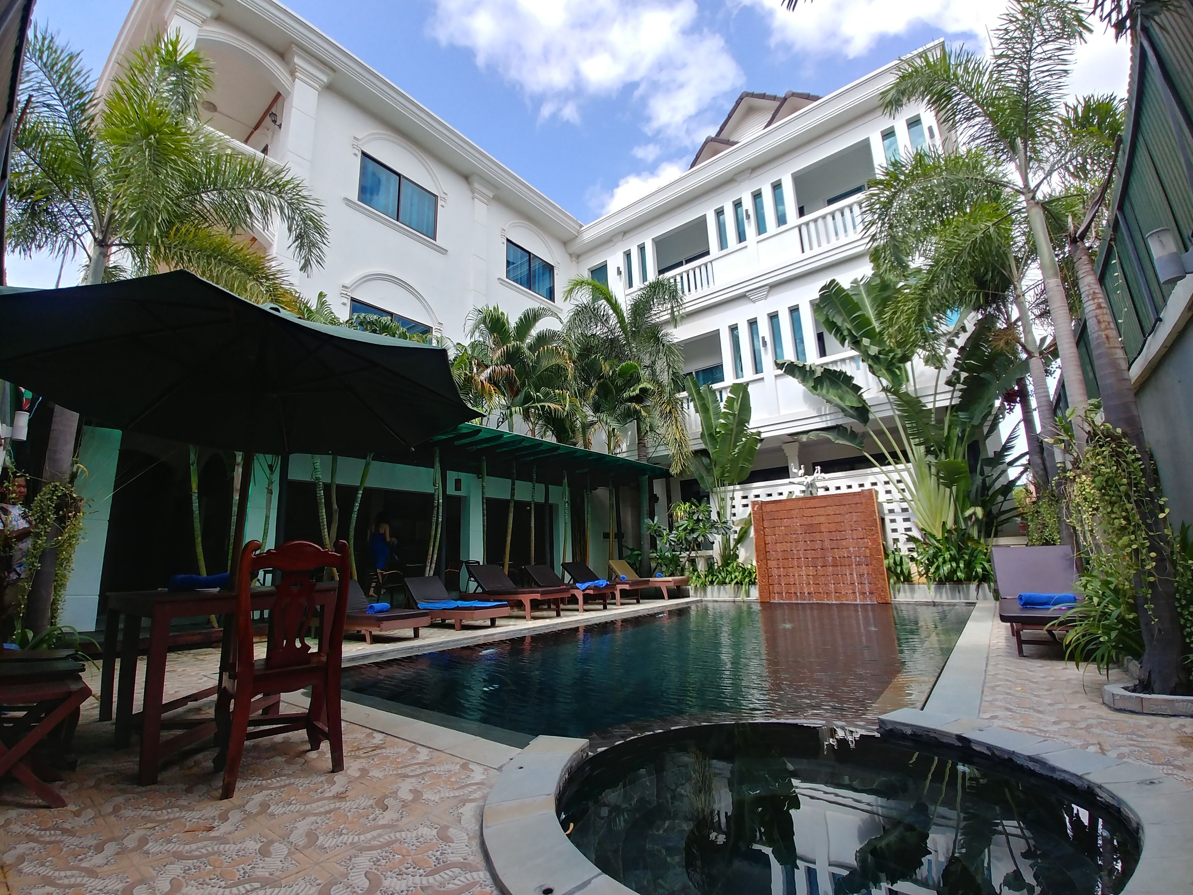 A pool area with lounge chairs and umbrellas, surrounded by lush greenery and modern buildings.