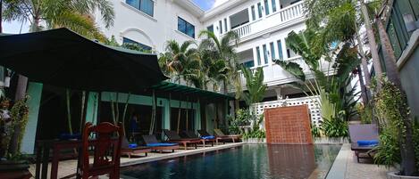 A pool area with lounge chairs and umbrellas, surrounded by lush greenery and modern buildings.