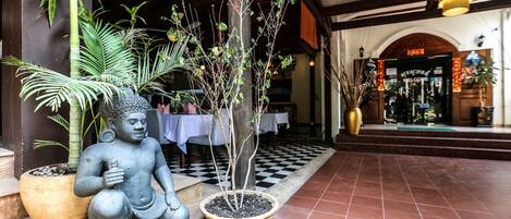 A statue of a seated figure next to a potted plant, with a checkered floor and a building entrance in the background.